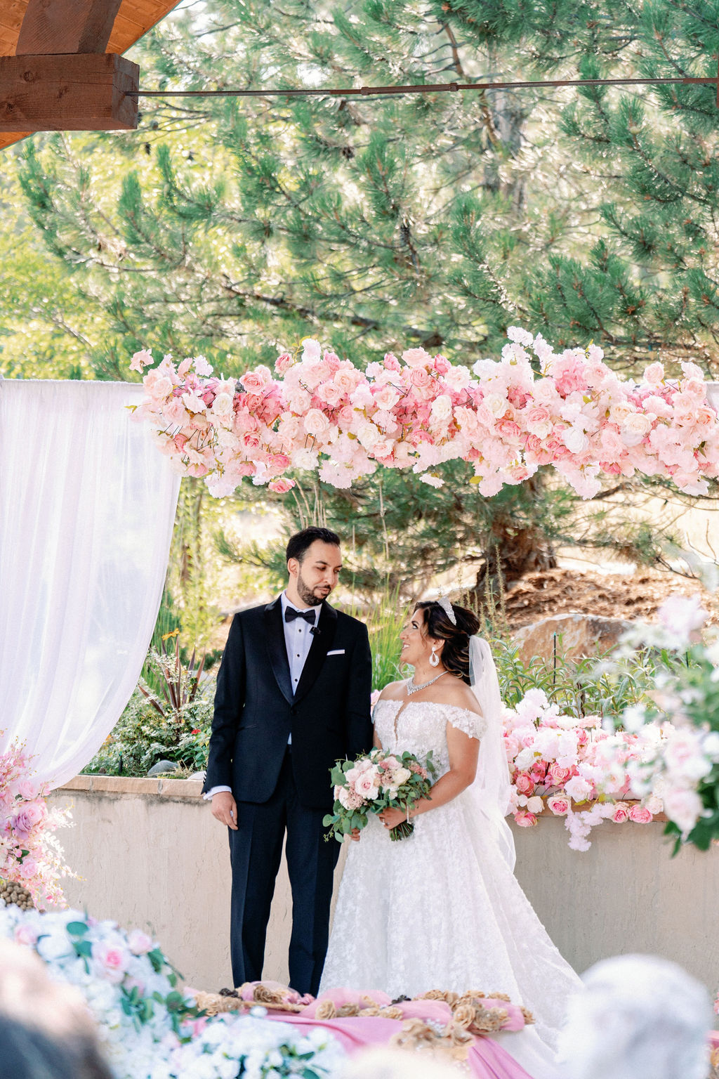 Newlyweds stand smiling at each other under an arch of pink roses