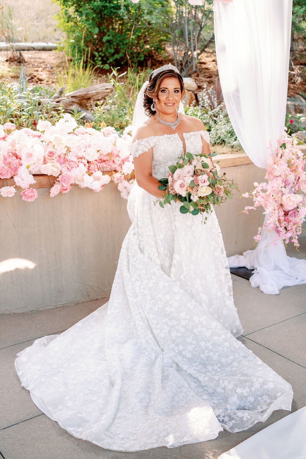 A bride smiles while holding her bouquet in her lace gown surrounded by pink roses at Cielo At Castle Pines