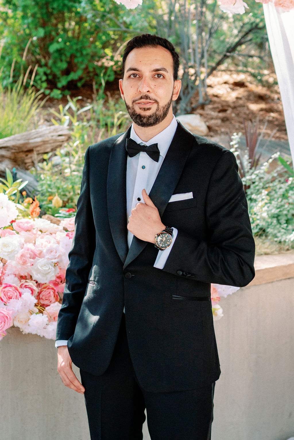 A groom holds his lapel while standing in the gardens with pink roses on the wall