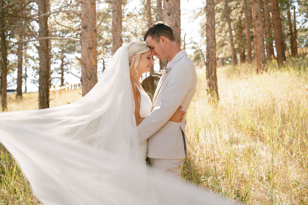 A bride and groom touch foreheads in the forst at sunset as the veil flows around them at one of the Boulder wedding venues