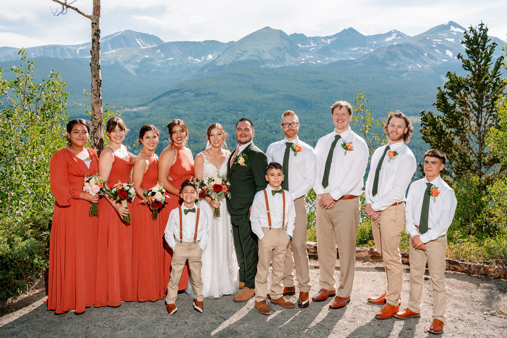 A bride and groom stand with their wedding party smiling with hands crossed at a mountain overlook