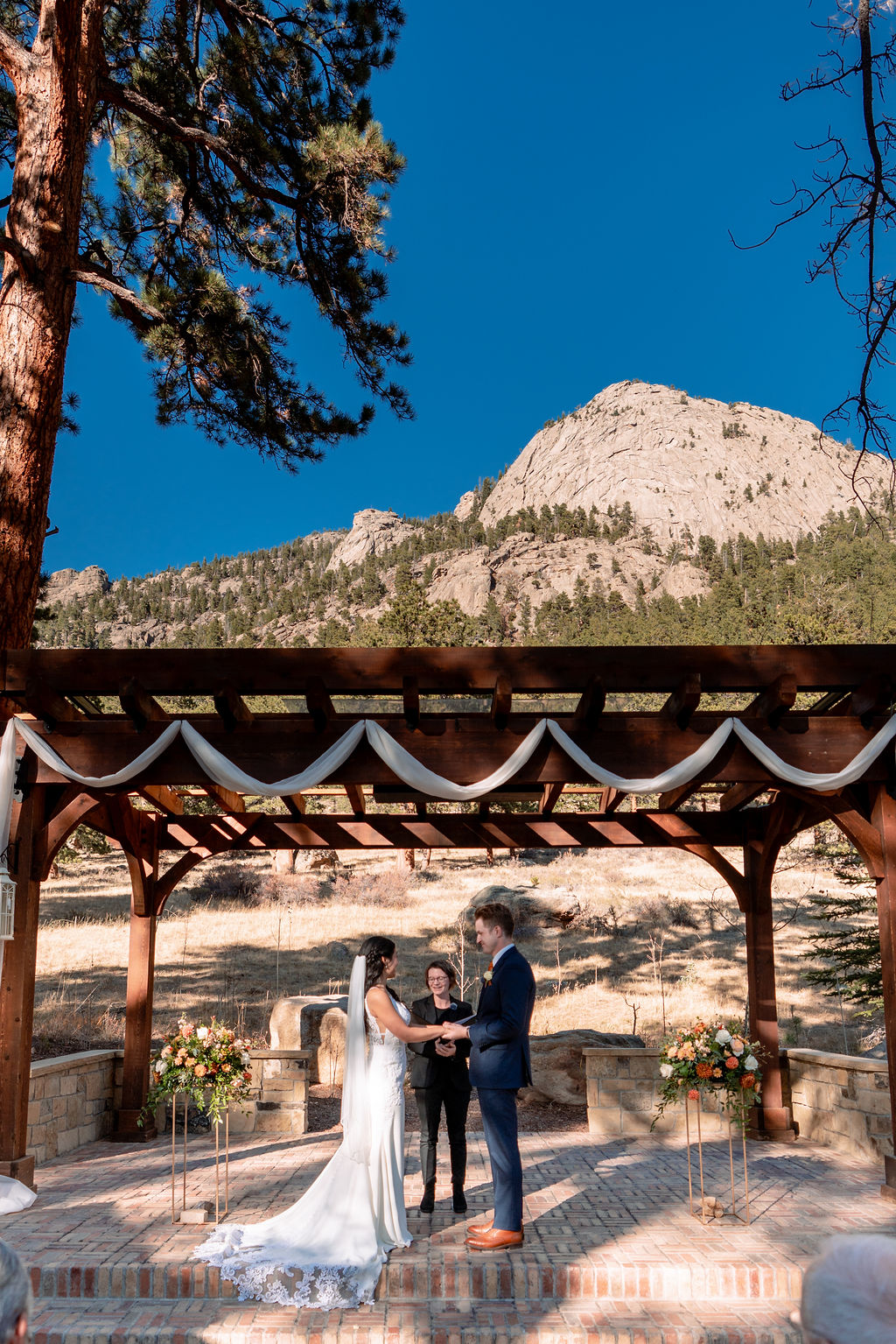 A peak at the bride and groom under a pergola during their Della Terra Wedding ceremony with mountain backdrop