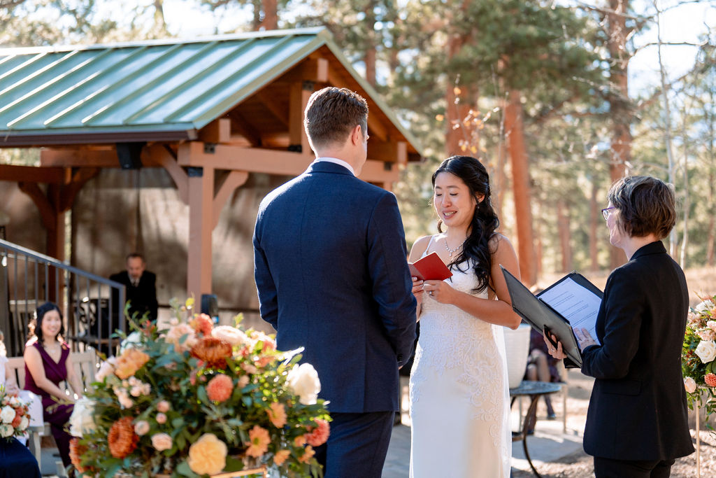 A bride reads from a red notebook to her groom during their outdoor ceremony