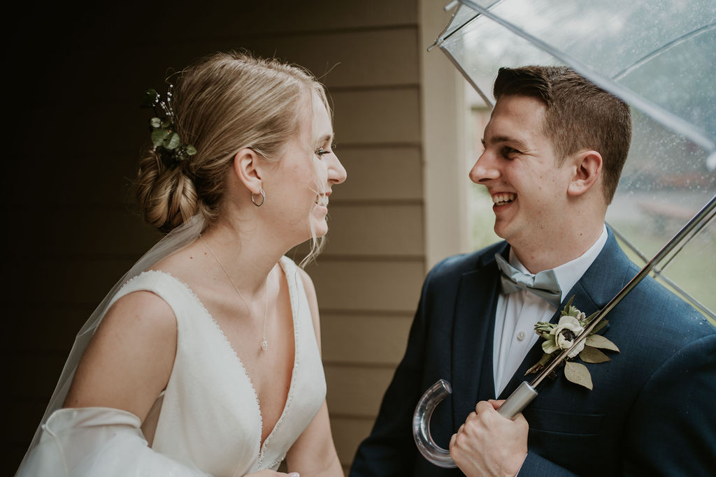 Newlyweds laugh together while hiding under a clear umbrella