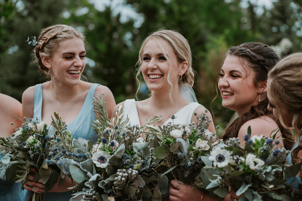 A bride laughs while standing with her bridesmaids holding their bouquets outside
