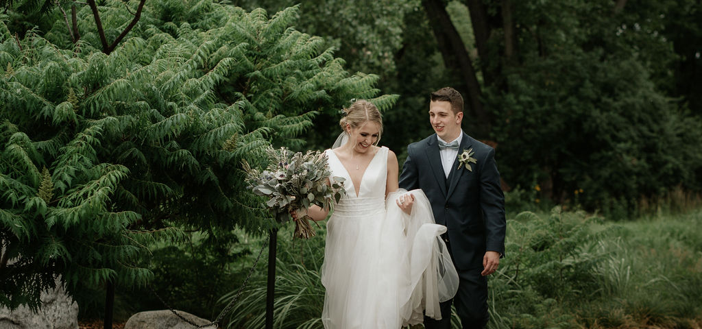 A bride and groom walk through the gardens of hudson gardens wedding venue while carrying the veil and train
