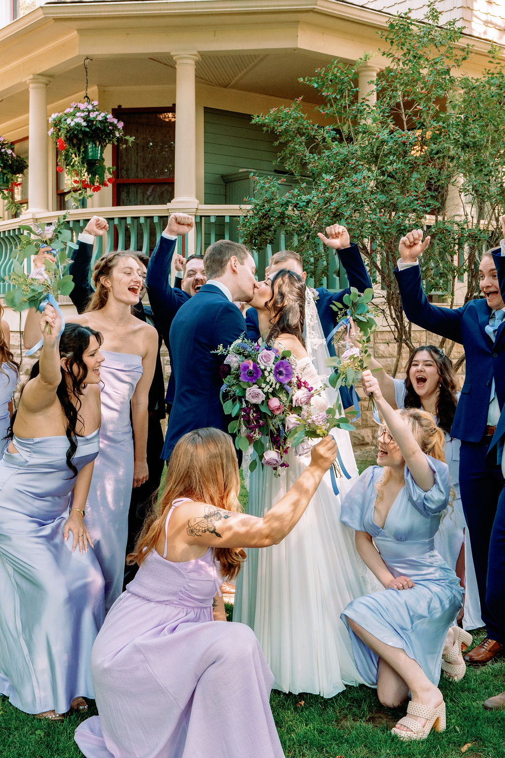 Newlyweds kiss in the lawn while their wedding party surrounds them cheering