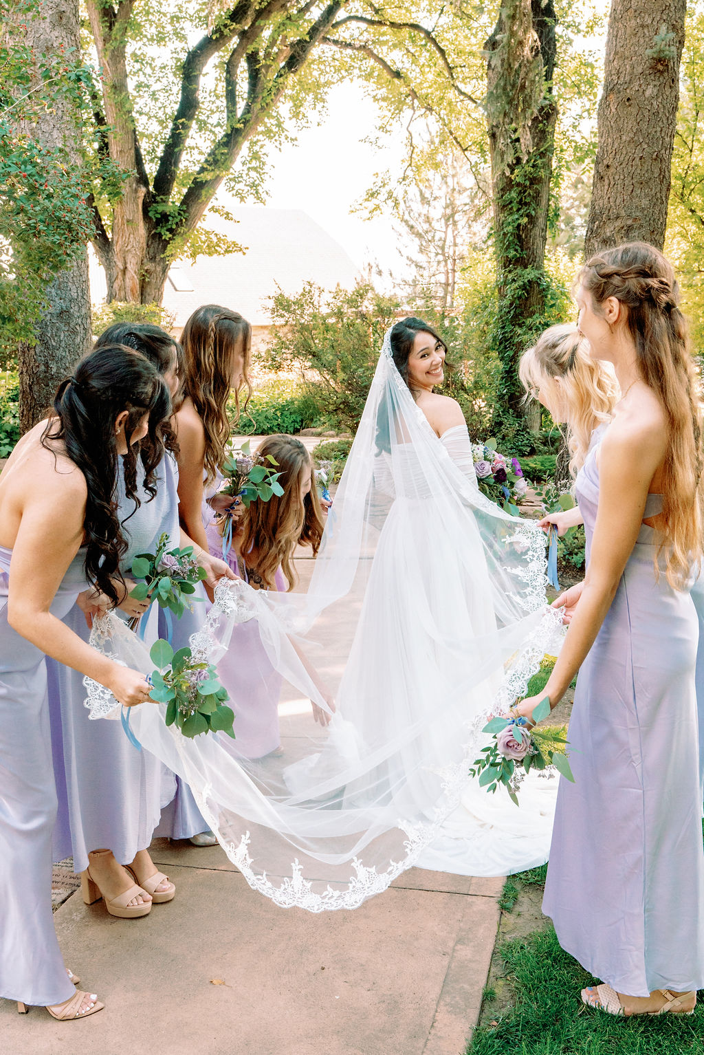 A bride smiles over her shoulder as her bridesmaids in purple hold her veil at Tapestry House By Wedgewood Weddings