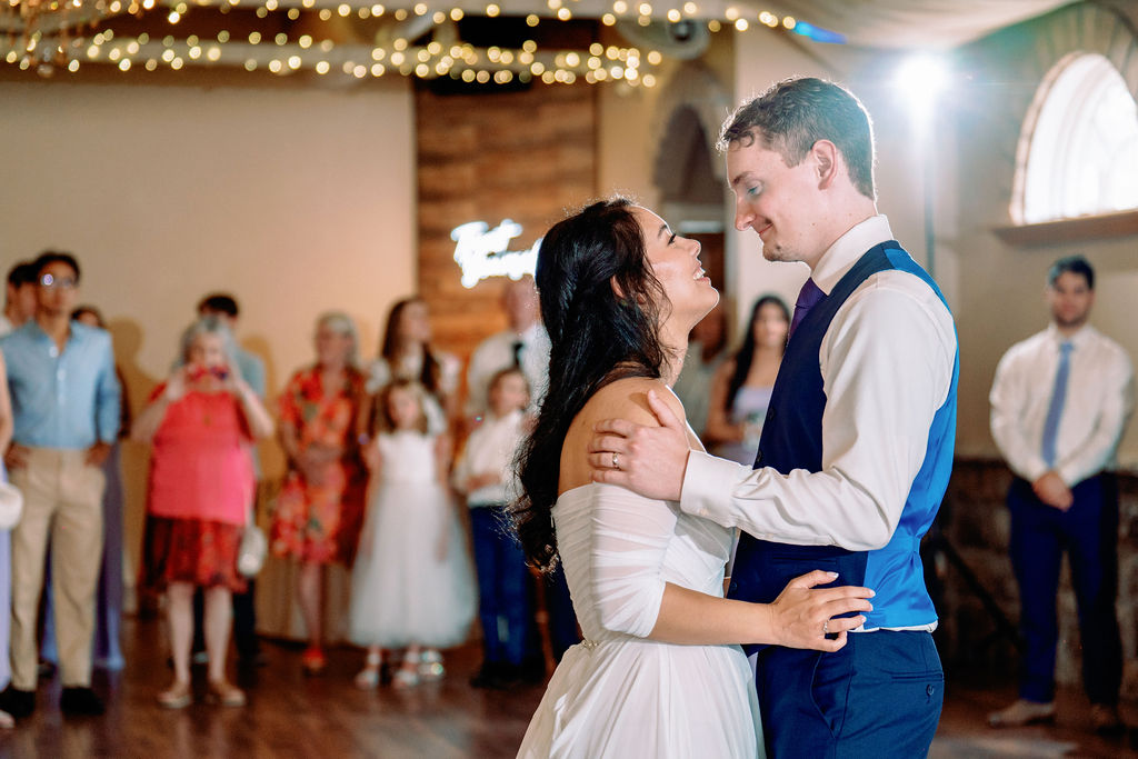 A bride and groom share their first dance while smiling at each other under market lights as guests look on at Tapestry House By Wedgewood Weddings