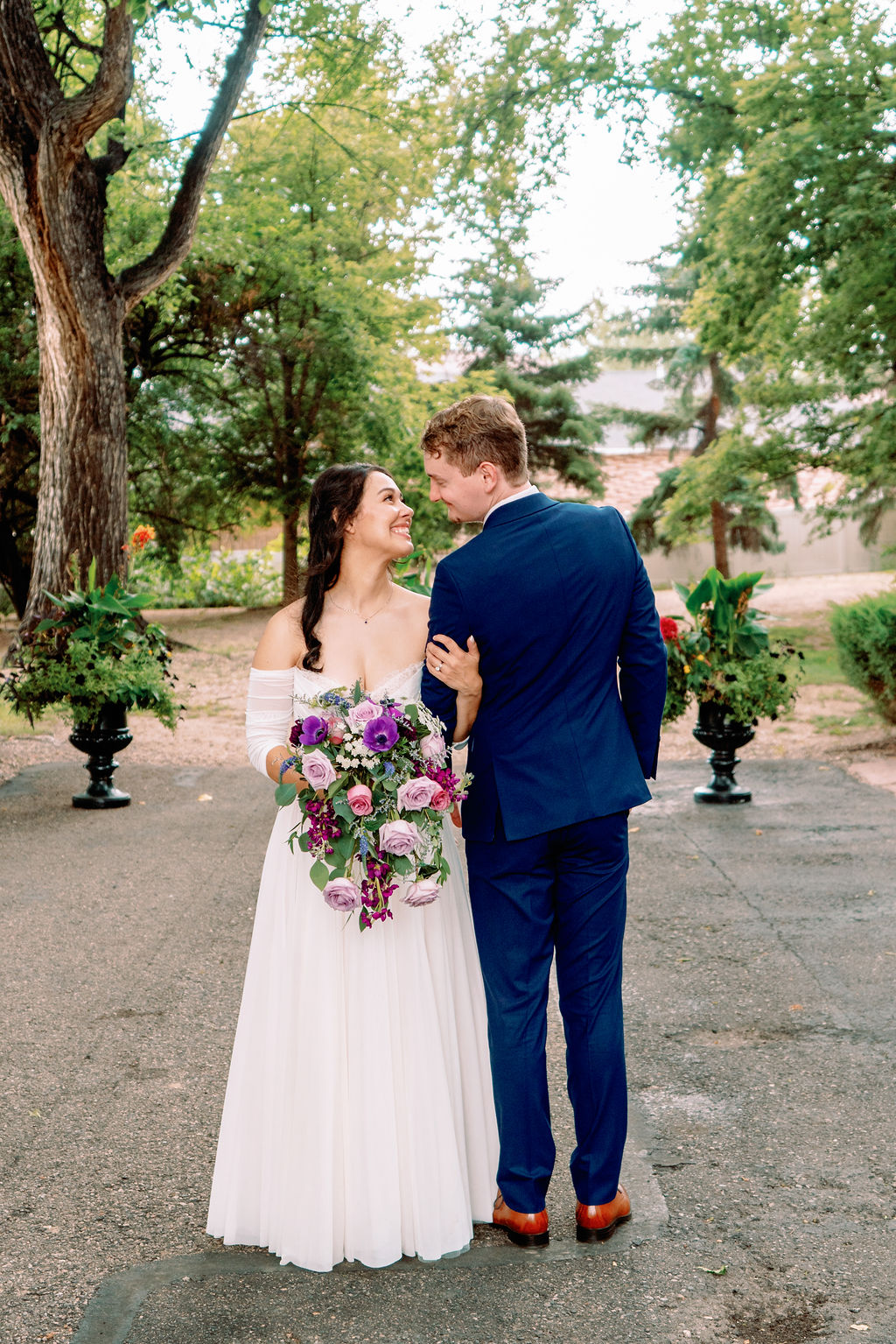 Newlyweds stand facing opposite directions while smiling at each other in a garden patio
