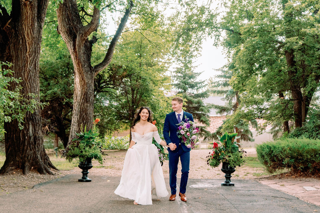 Newlyweds hold hands while walking in the gardens as the groom holds the bright bouquet at Tapestry House By Wedgewood Weddings