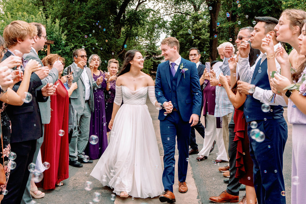 Happy newlyweds smile at each other while holding hands and walking through bubbles blows by their guests