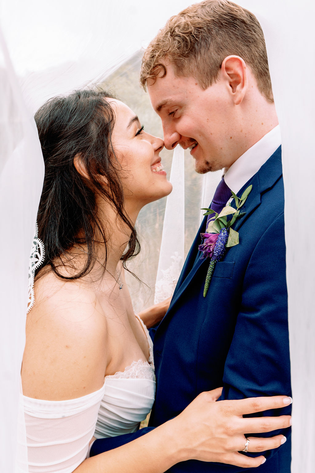 A bride and groom boop noses while smiling at each other and hiding under the veil
