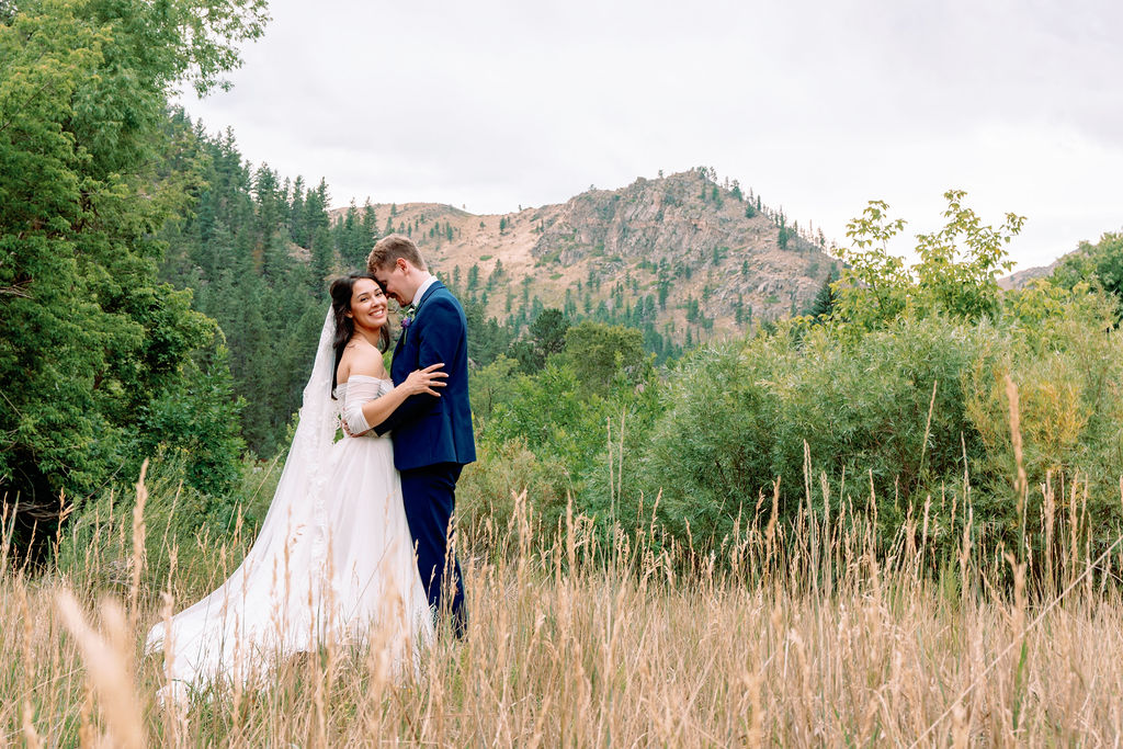 A bride smiles big while hugging her groom in his blue suit in tall golden grass in the Rocky Mountains