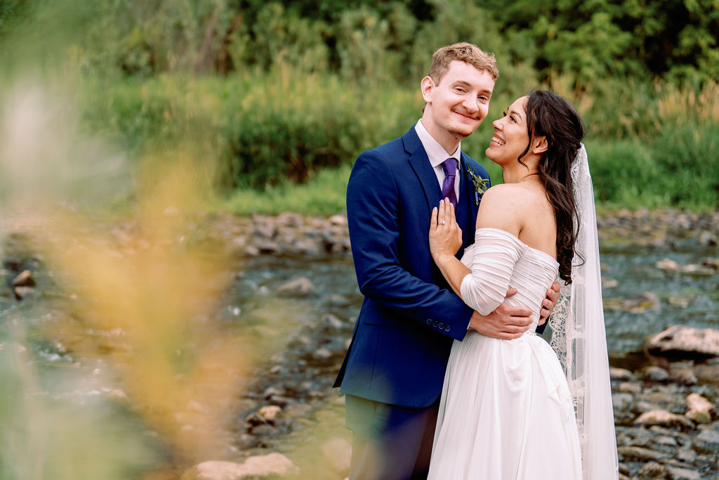 A bride smiles up to her groom as they hug by a creek