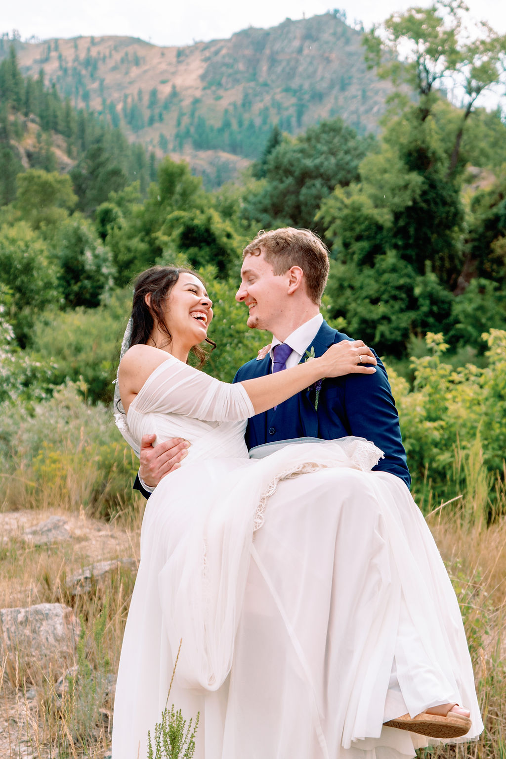 A smiling groom in a blue suit carries his laughing bride through a forest trail