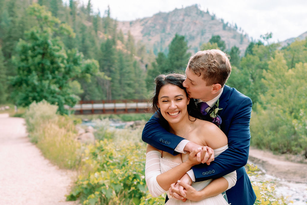 A groom hugs and kisses the temple of his bride while exploring a riverside path at Tapestry House By Wedgewood Weddings