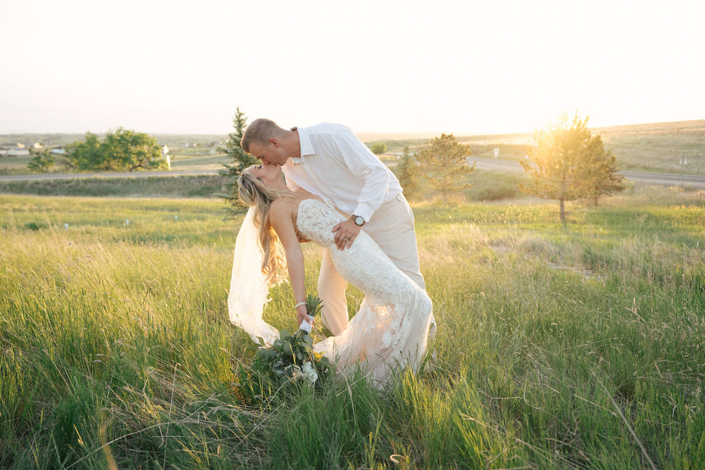 A groom dips his bride for a kiss in the tall grass at sunset