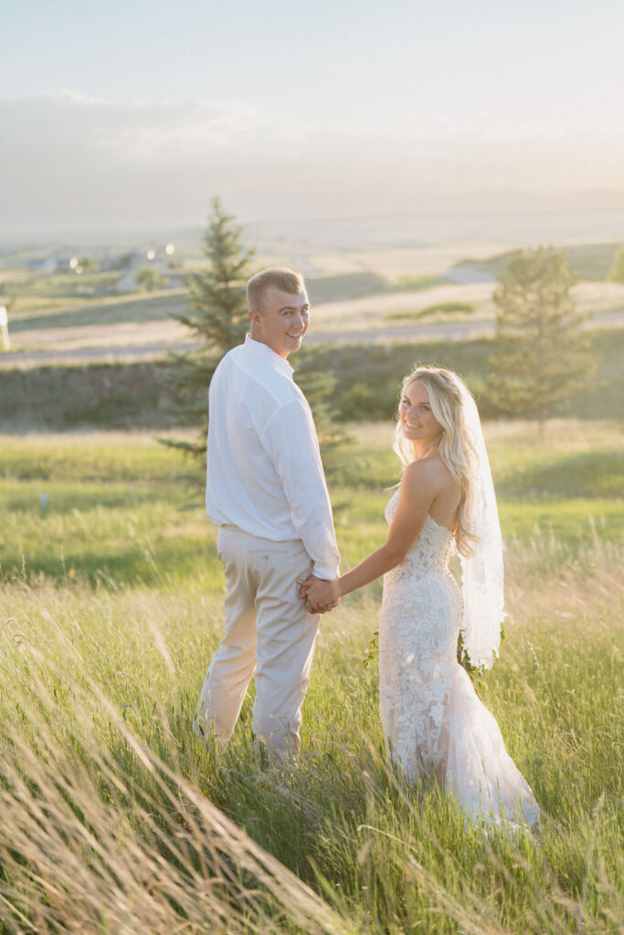 Newlyweds hold hands and smile over their shoulder while admiring sunset on a hillside of tall grass at wedding venues near Denver