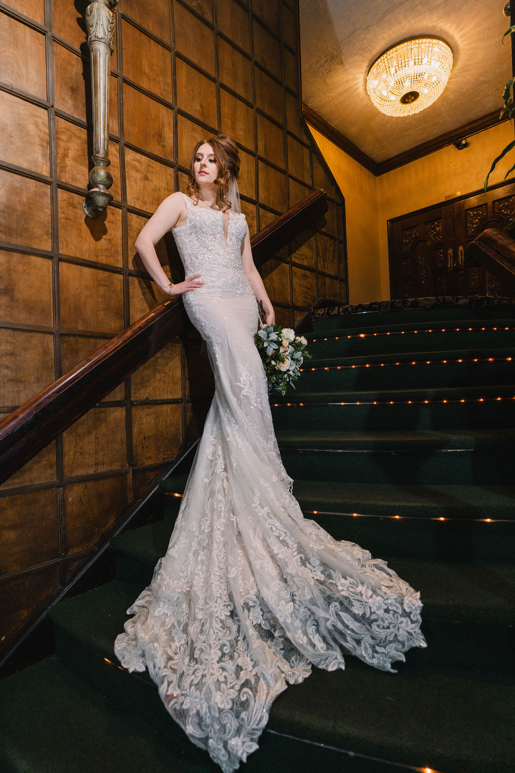 A bride leans on the wooden railing of a staircase with a hand on her hip and train flowing around her at Wellshire Event Center