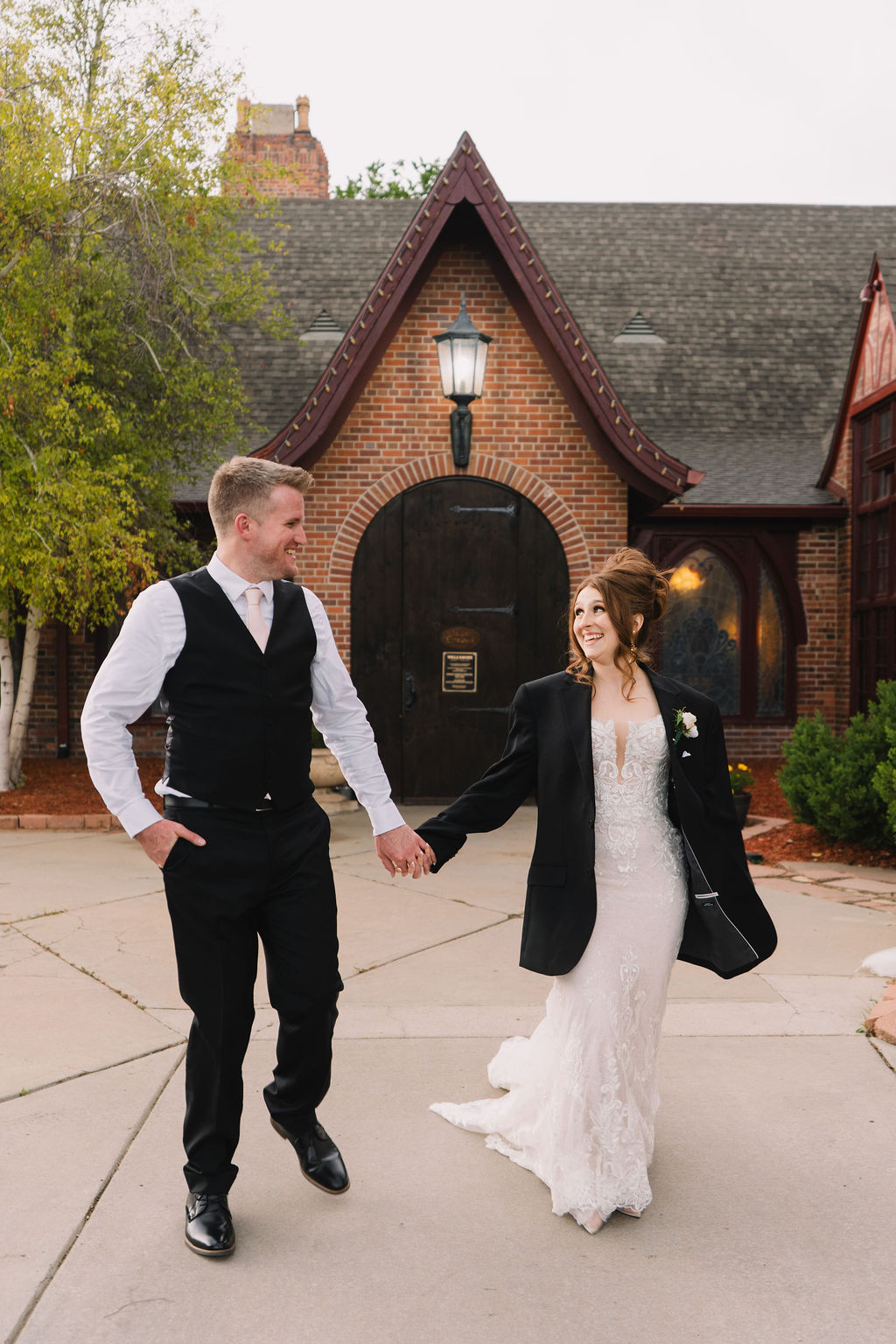 A bride wearing her groom's jacket walks with him holding hands in the driveway of one of the wedding venues near Denver