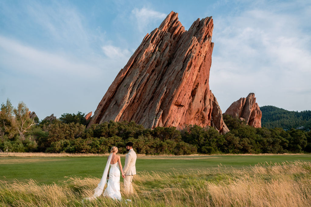 A. bride and groom hold hands while chatting in tall golden grass by an astonishing rock formation