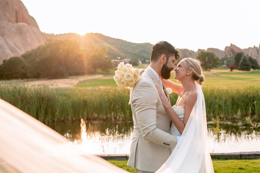 Newlyweds embrace for a kiss by a pond at sunset as the veil wraps around them at one of the beautiful wedding venues near Denver