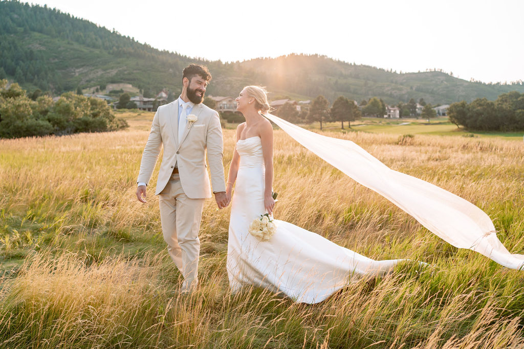 Newlyweds smile at each other while walking through a windy field at sunset