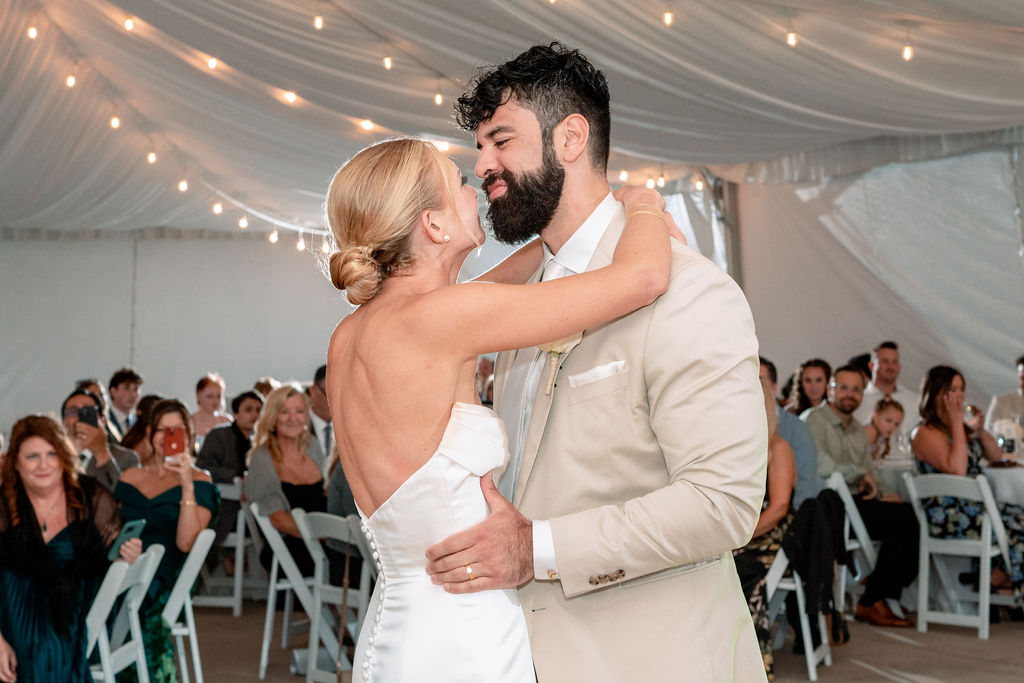 Newlyweds dance close and lean in for a kiss under a tent as guests watch