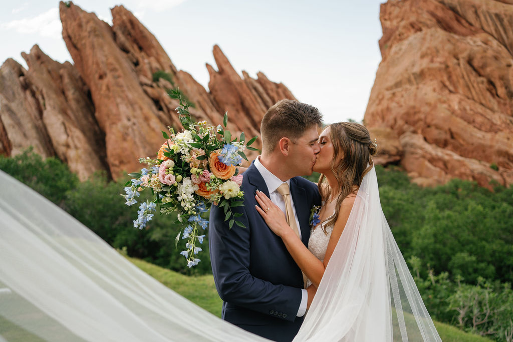 Newlyweds kiss while the veil flows around them and holding a large vibrant bouquet at one of the wedding venues near Denver with red rocks