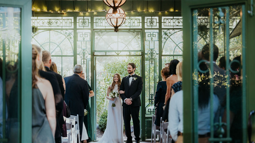 A bride and groom smile together at the end of their ceremony in a greenhouse as guests stand for their exit