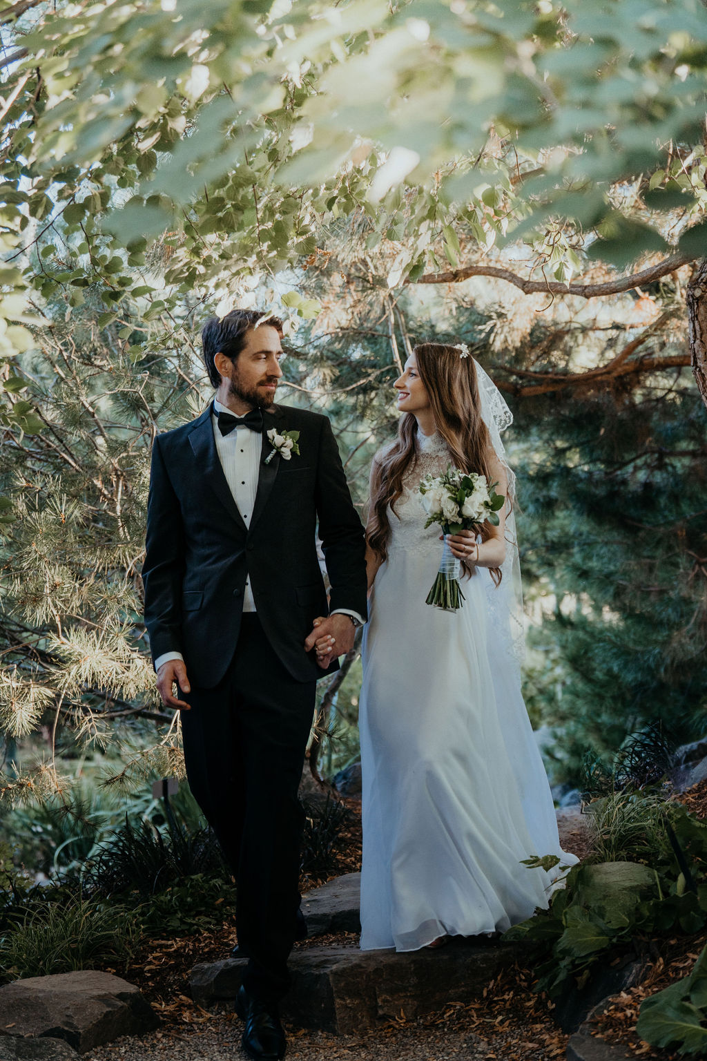 A bride and groom smile at each other while holding hands on a quiet garden trail at sunset at one of the wedding venues near Denver