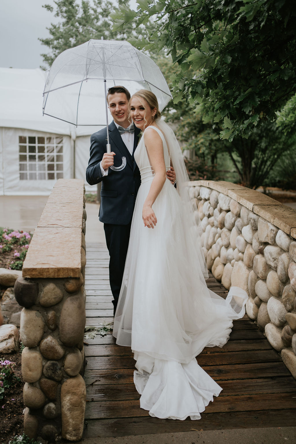 A bride and groom laugh while walking over a stone and wood bridge at one of the wedding venues near Denver under a clear umbrella