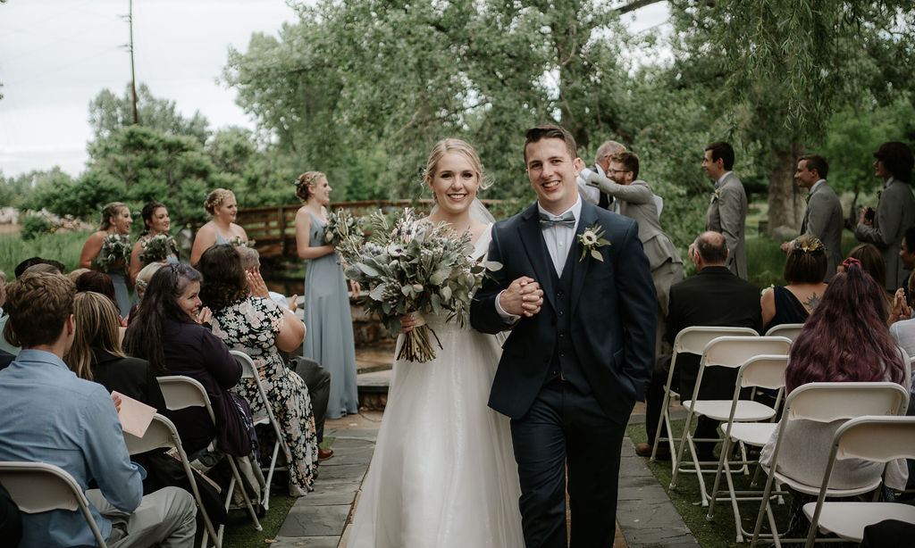 Newlyweds smile while holding hands and large bouquet walking up the aisle ending their ceremony at one of the wedding venues near Denver