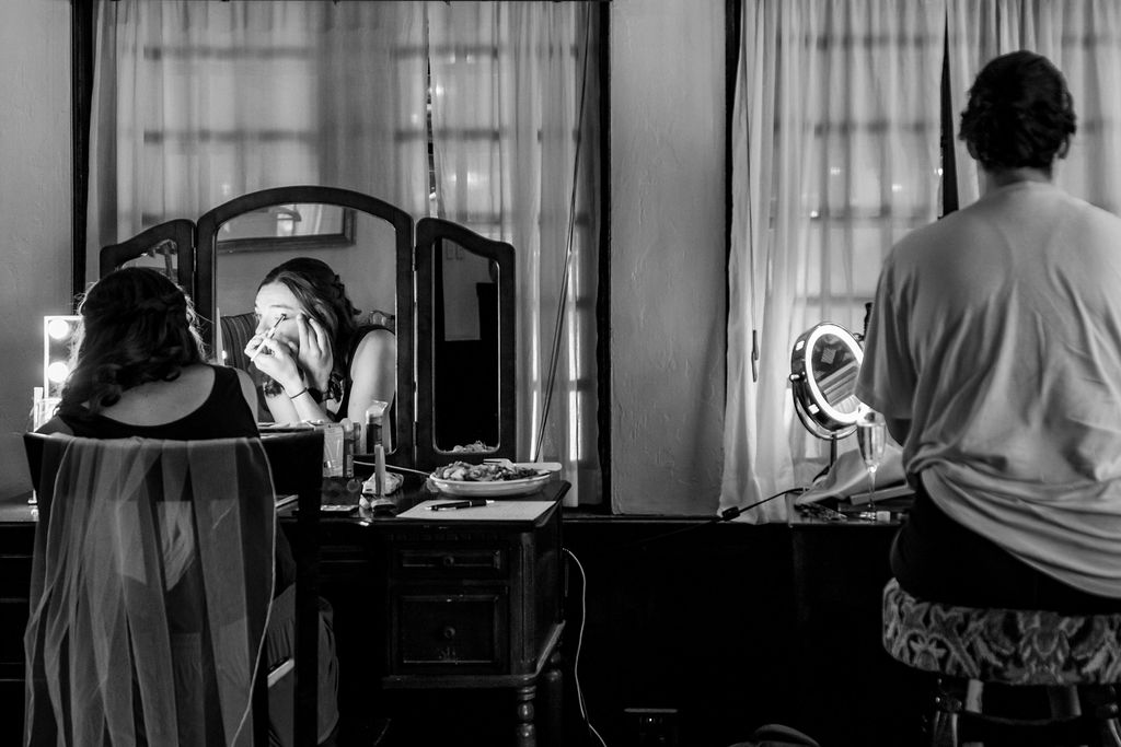 A bride applies makeup in a three panel mirror in black and white