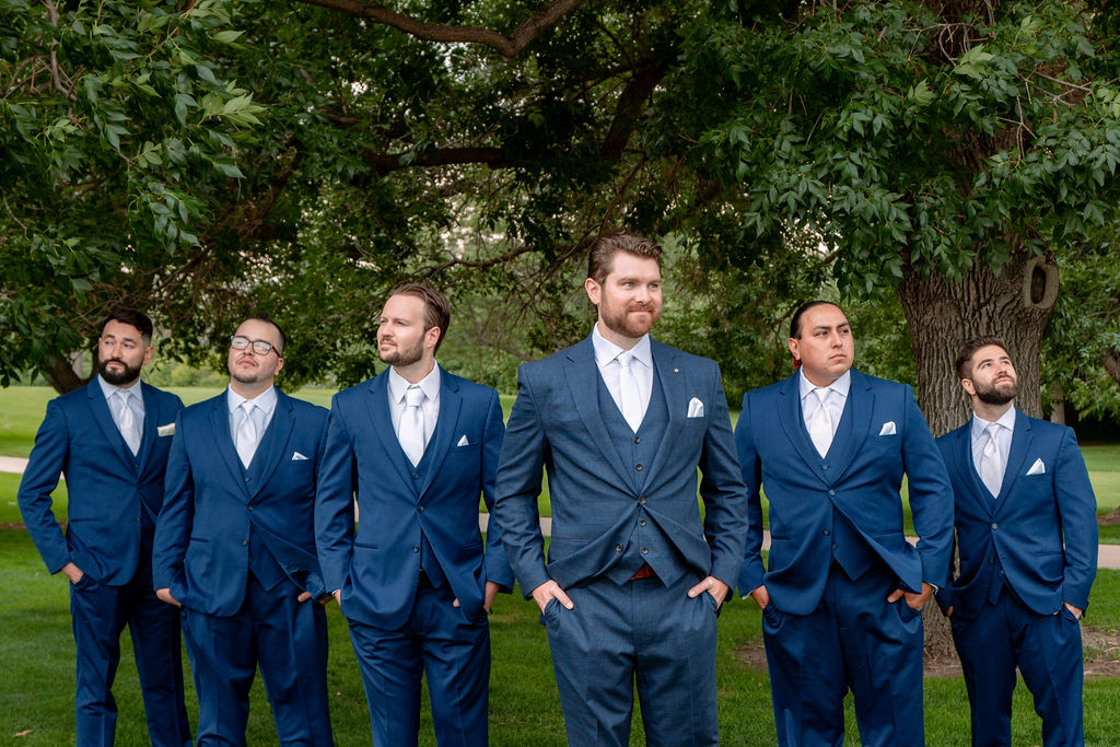 A groom in a blue suit stands with hands in his pockets with his five groomsmen in matching suits